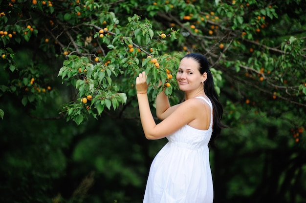 Pregnant girl in a white sundress on the background of apricot trees. Pregnant woman smiling