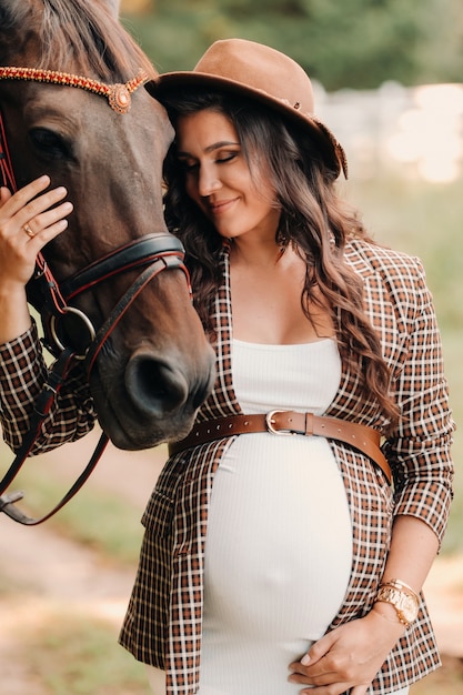 Pregnant girl with a big belly in a hat next to horses in the forest in nature