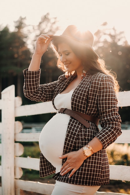 日没時の自然の中で馬の囲いの近くの帽子に大きな腹を持つ妊婦。馬と茶色のドレスを着たスタイリッシュな妊婦。