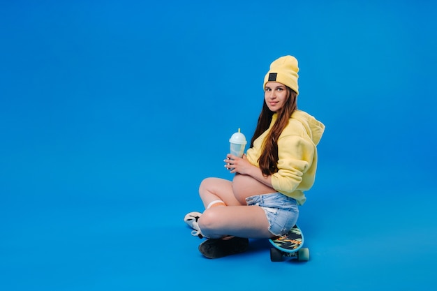 A pregnant girl in yellow clothes with a glass of juice sits on a skateboard on a blue background.