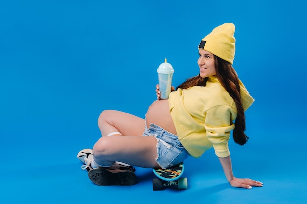 A pregnant girl in yellow clothes with a glass of juice sits on a skateboard on a blue background.