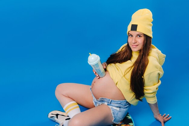 A pregnant girl in yellow clothes with a glass of juice sits on a skateboard on a blue background.