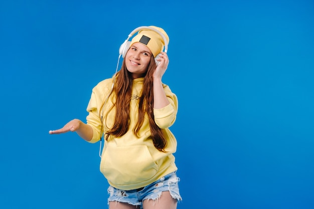 A pregnant girl in a yellow jacket and headphones stands on a blue background.