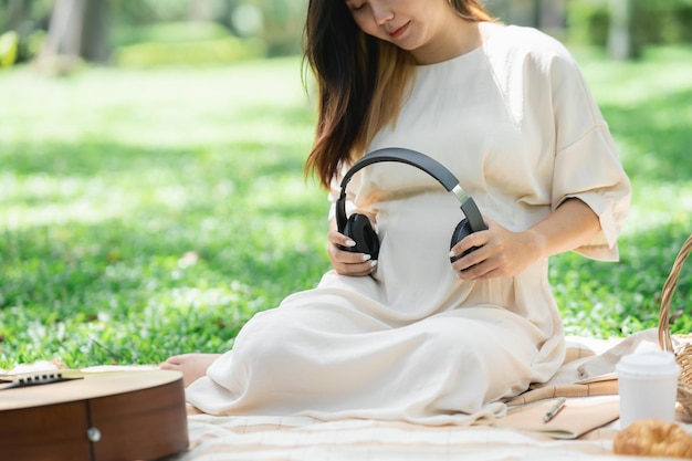 Pregnant happy asian woman putting headphones on her belly to listen music while sitting on picnic blanket on lawn at the garden park under the tree Family mother mom pregnant healthy concept