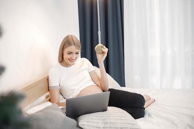 Pregnant lady lie on a bed in room and holding an apple while\
using a laptop