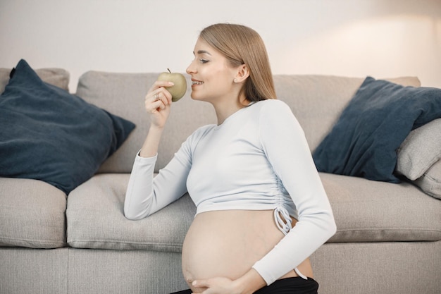 Pregnant lady sitting near a sofa in living room holding an\
apple