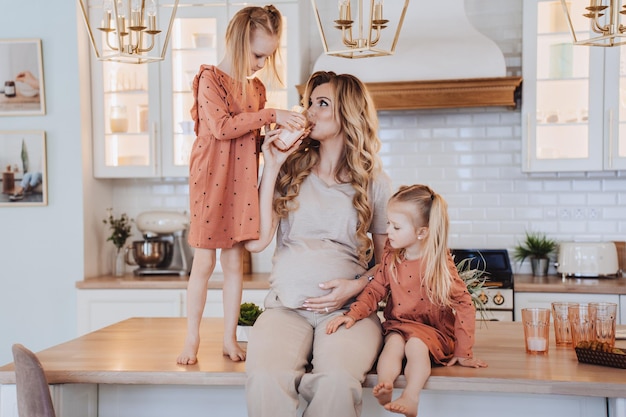 Pregnant mom sitting on a kitchen table with little daughters dressed in beige casual clothes