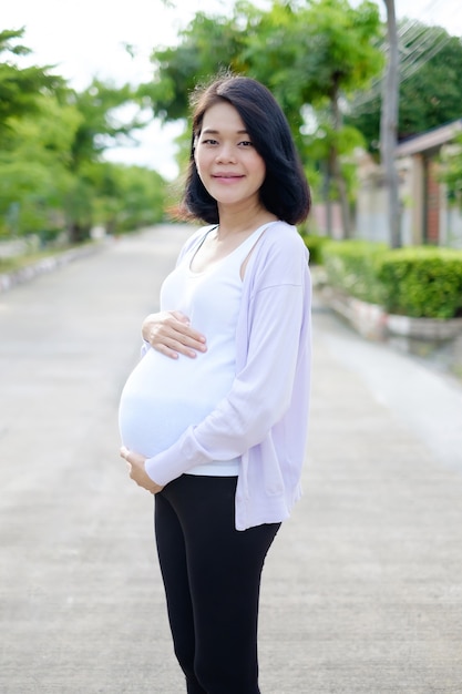 A pregnant mother in casual clothes is smiling on the road against a green tree.