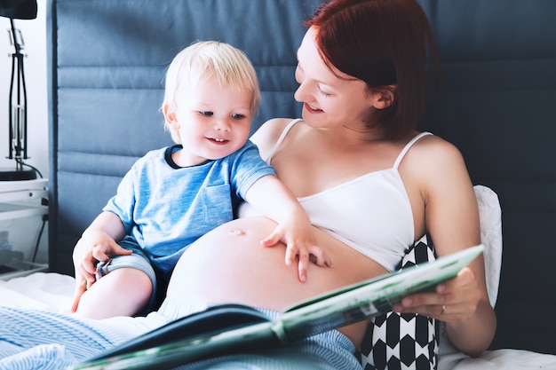 Pregnant mother and son together in bed at home Little child boy looking at her mother tummy