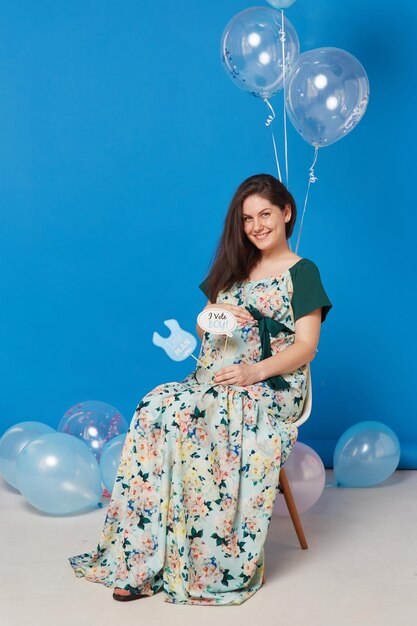 Pregnant smiling brunette young woman sitting on the chair with balloons isolated blue background