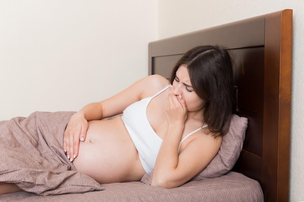 吐き気に苦しむ妊娠中の妊娠症状期待中毒症ベッドの親の概念に座っている若い嘔吐女性