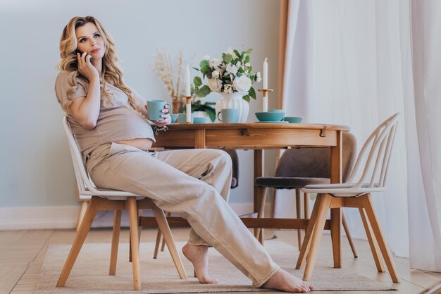 Pregnant swedish woman in beige tshirt and pants holding up talking by phone sits at table home