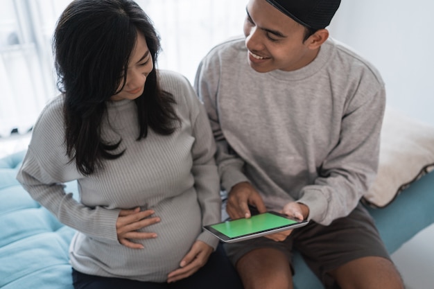 A pregnant wife and husband sitting while holding a digital tablet