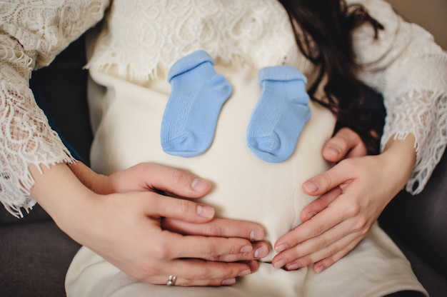 A pregnant wife, man holds socks her arms on around round stomach. View of the stomach pregnant wife. waiting a baby. Happy portrait, concept of a family holiday. Close up.