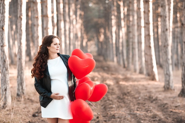 Pregnant woman 30-35 year old holding heart shape red balloons standing in forest