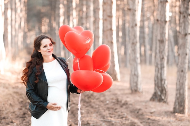 Pregnant woman 30-35 year old holding heart shape red balloons standing in forest