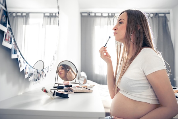 A pregnant woman applies makeup at home in front of a mirror