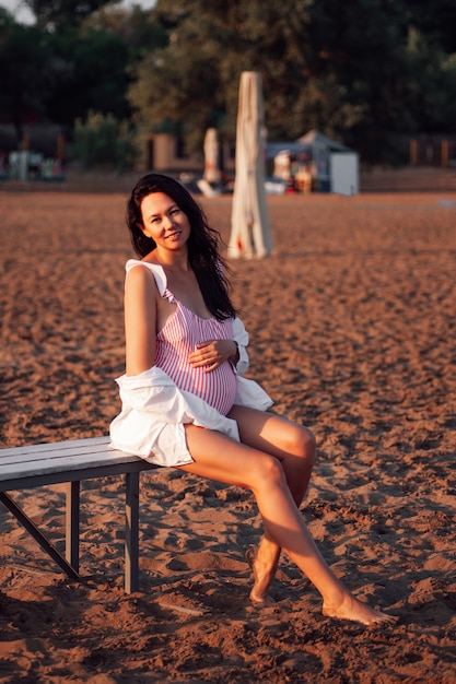 A pregnant woman on the beach a happy beautiful pregnant woman in a pink swimsuit and a white shirt ...