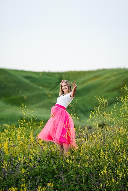 아름다운 분홍색 드레스를 입은 임산부가 꽃밭을 걷고 임신을 즐깁니다.