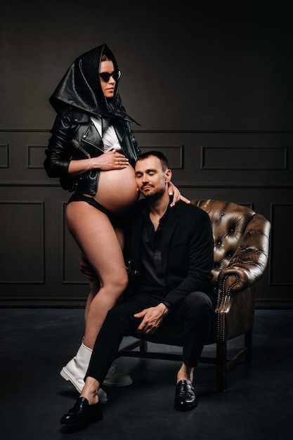 A pregnant woman in black clothes and a headscarf and a man in a suit in a studio on a dark background.