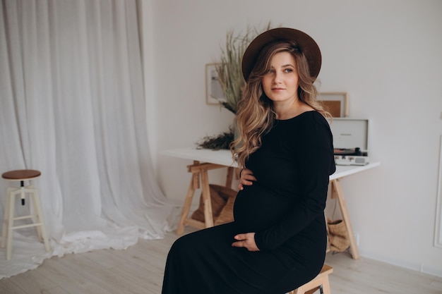 Беременная женщина в черном платье сидит на стуле в студии на белом фоне