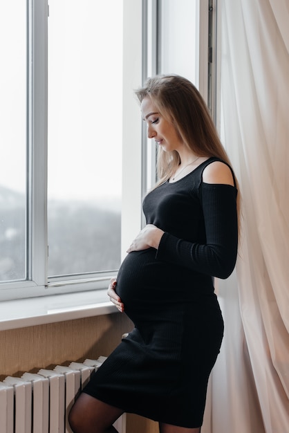 Беременная женщина в черном платье стоит у окна