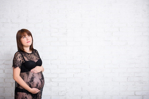 Pregnant woman in black lace dress thinking or dreaming about something over white brick wall