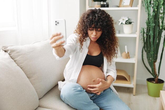 Photo pregnant woman blogger sits on the couch at home and takes pictures of herself on the phone selfie