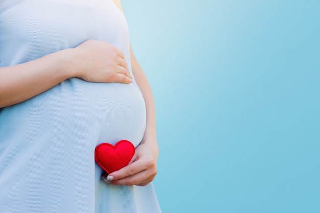 A pregnant woman in a blue dress holds in her hands a red heart on blue. Pregnancy concept