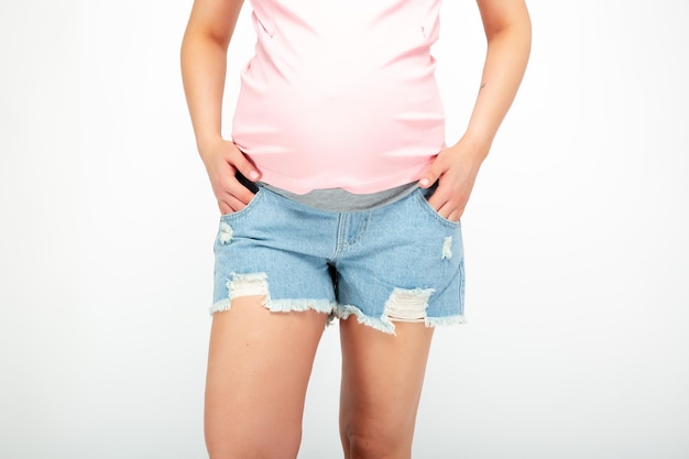 A pregnant woman in blue shorts and a pink shirt stands with her hands in her pockets.
