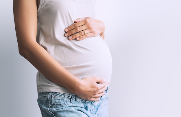 Pregnant woman in casual clothing holds hands on belly on white background
