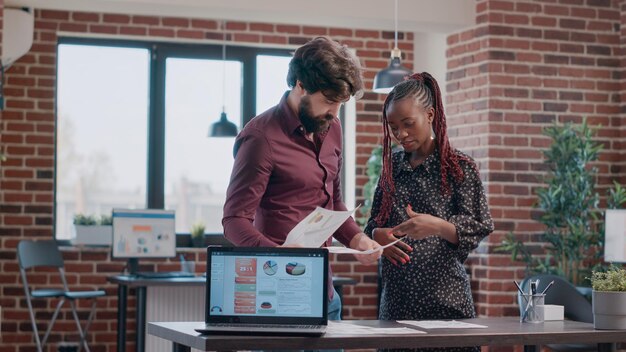 Pregnant woman and colleague brainstorming ideas for project planning in office. Business man holding laptop with data while person expecting child analyzing marketing strategy.