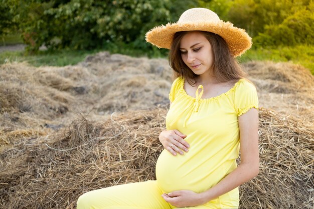 A pregnant woman in dark clothes and a hat sits in a field on straw in summer