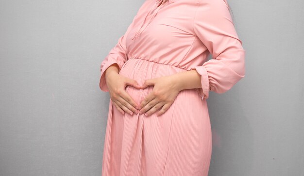 Pregnant woman in dress holds hands on belly on grey.