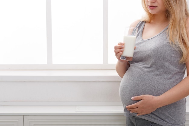 Pregnant woman drinking glass of milk copy space. Young unrecognizable expectant blonde enjoying healthy drink, standing near window. Healthcare, nutrition, vitamins, pregnancy concept