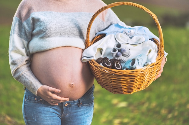 Pregnant woman expecting a baby, posing outdoors with a wicker basket with clothes for her newborn.