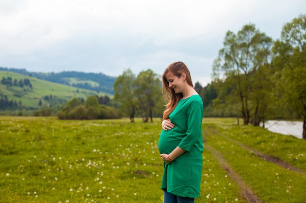 Pregnant woman in a green tunic is breathing clear air on the background of nature with green trees