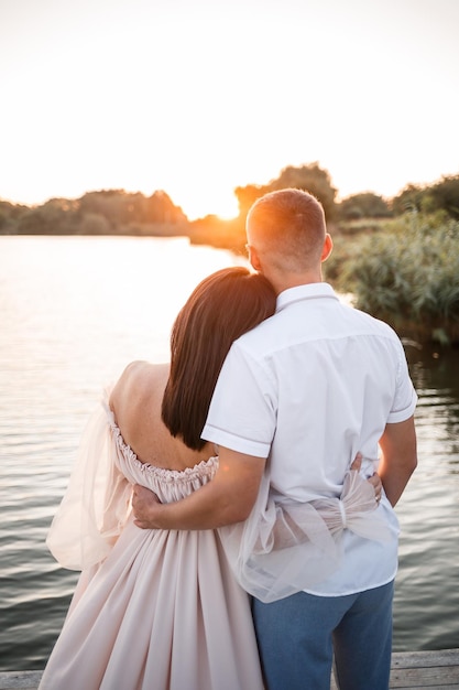 Una donna incinta e suo marito stanno sul molo e si guardano un bellissimo fiume e al tramonto i futuri genitori trascorrono del tempo all'aperto una bellissima giovane madre