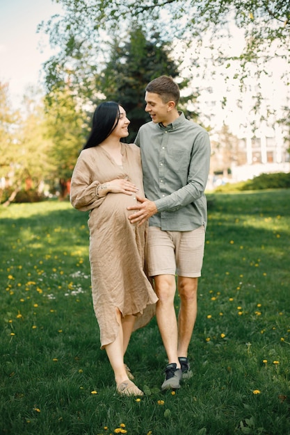 Pregnant woman and her husband standing in a park on a grass and hugging