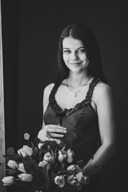 Pregnant woman hold bouquet of flowers in hands girl embraces a round stomach belly standing near the window at home Maternity concept Close up nine months Baby Shower Black and white photo