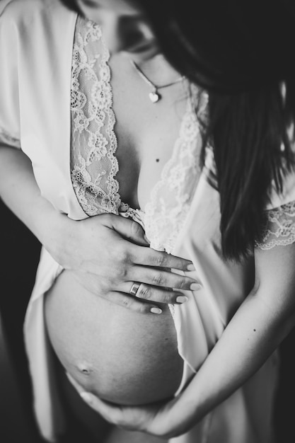 Donna incinta tenere le mani intorno allo stomaco ragazza abbraccia una pancia rotonda vicino alla finestra a casa concetto di maternità primo piano di nove mesi baby shower vista frontale foto in bianco e nero