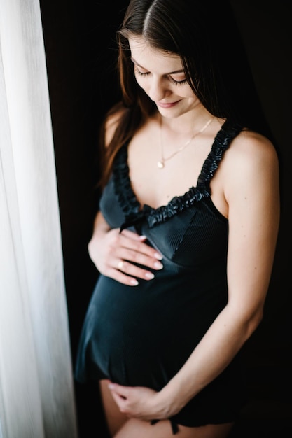 Pregnant Woman hold hands round stomach girl embraces a round belly near the window at home Standing sideways Maternity concept Close up nine months Baby Shower