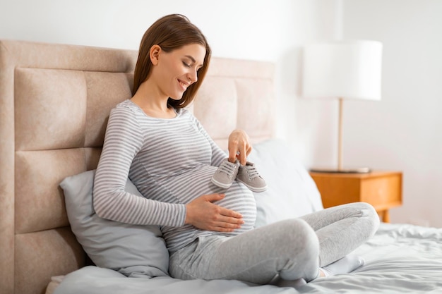 Pregnant woman holding baby shoes on bed