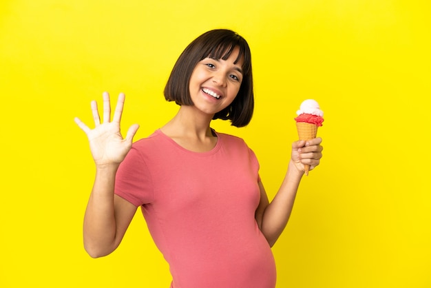노란색 배경에 격리된 코넷 아이스크림을 들고 있는 임산부는 손가락으로 다섯을 세고 있다