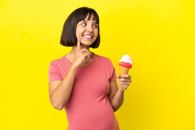 Pregnant woman holding a cornet ice cream isolated on yellow background thinking an idea while looking up