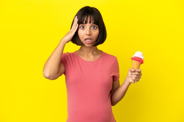 Pregnant woman holding a cornet ice cream isolated on yellow background with surprise expression