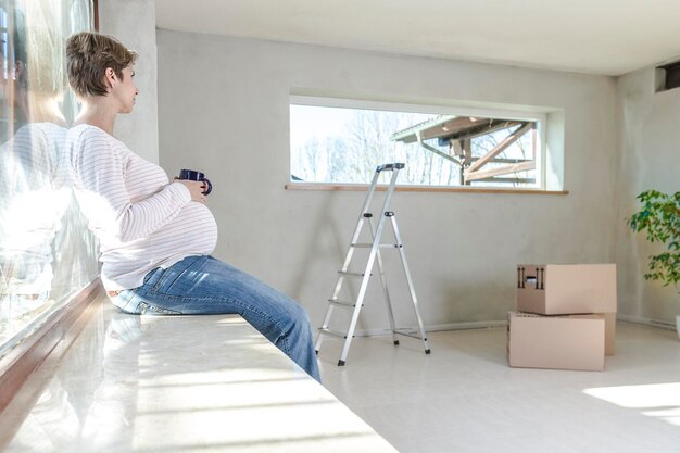 家の窓のそばに座っている中カップを握っている妊婦