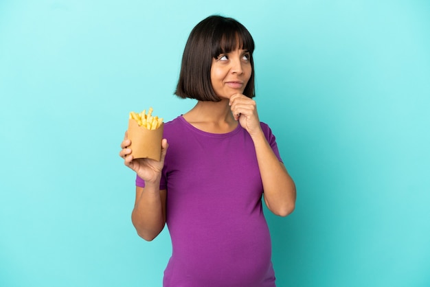 Pregnant woman holding fried chips over isolated background having doubts and thinking