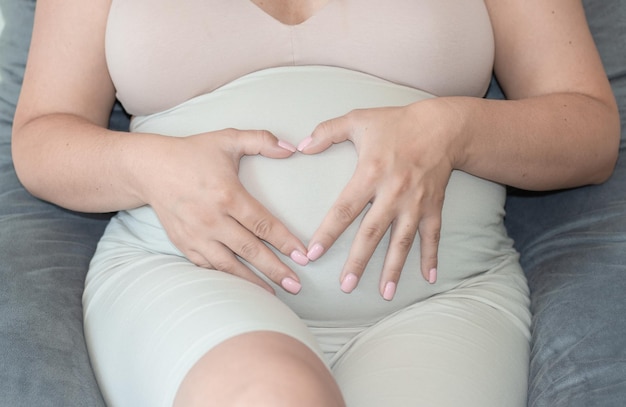 беременная женщина держит форму сердца обеими руками от пальцев над большим животом. яркое светлое фото