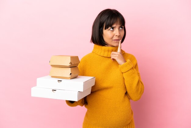 Pregnant woman holding pizzas and burgers isolated on pink background having doubts while looking up